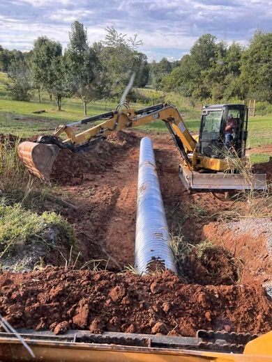 Excavator laying a long, corrugated metal pipe in a dirt trench in a field.