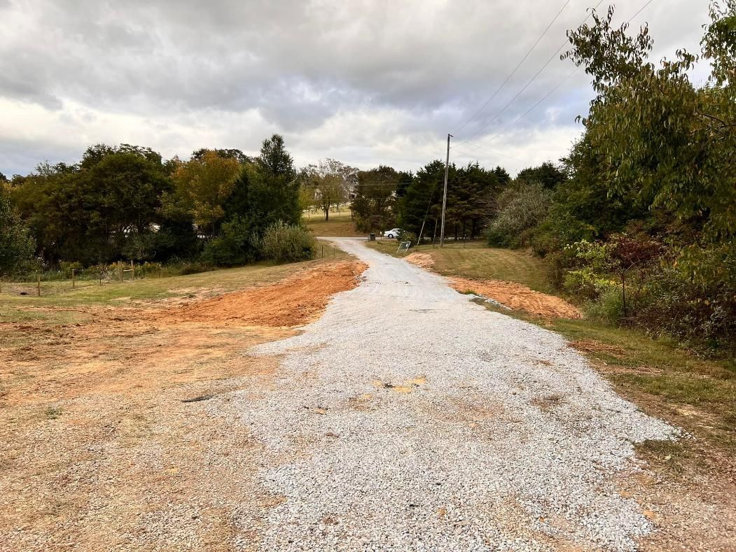Gravel driveway leading into the distance on a cloudy day, surrounded by brown dirt and green trees.