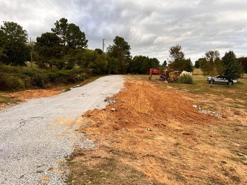 A gravel road under construction with dirt piles, an excavator, and a truck on a cloudy day.