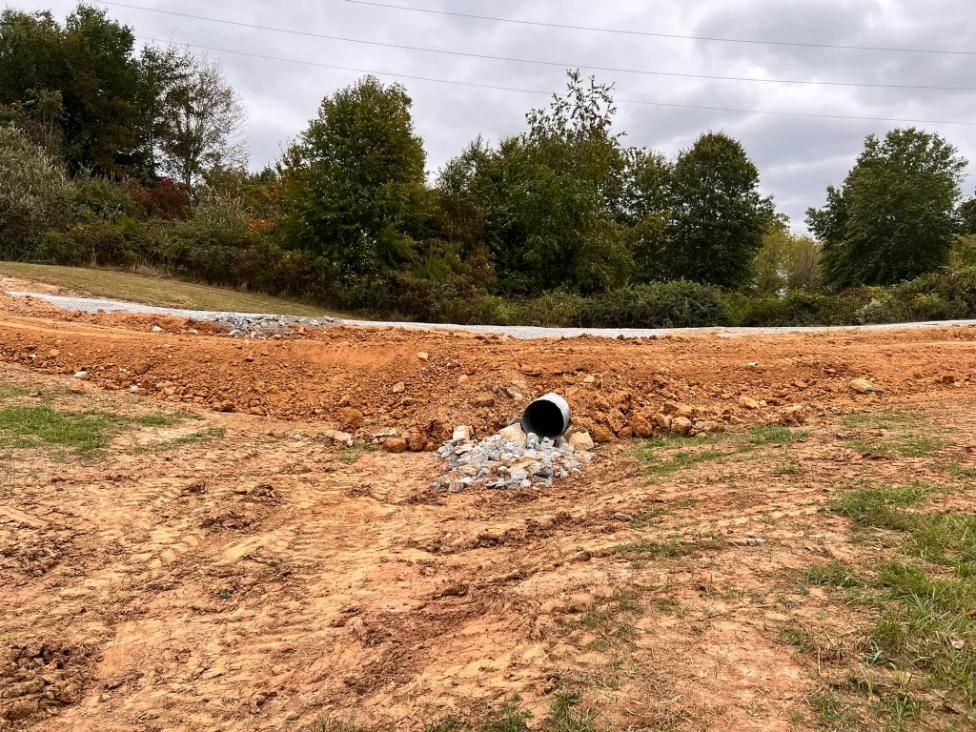 Dirt road with a drainage pipe leading to a rocky area, surrounded by brown earth and greenery under a cloudy sky.