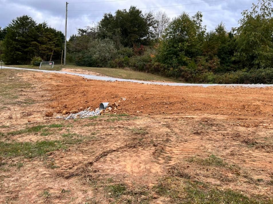 Dirt and gravel road construction in progress with a drain pipe visible, trees in background.