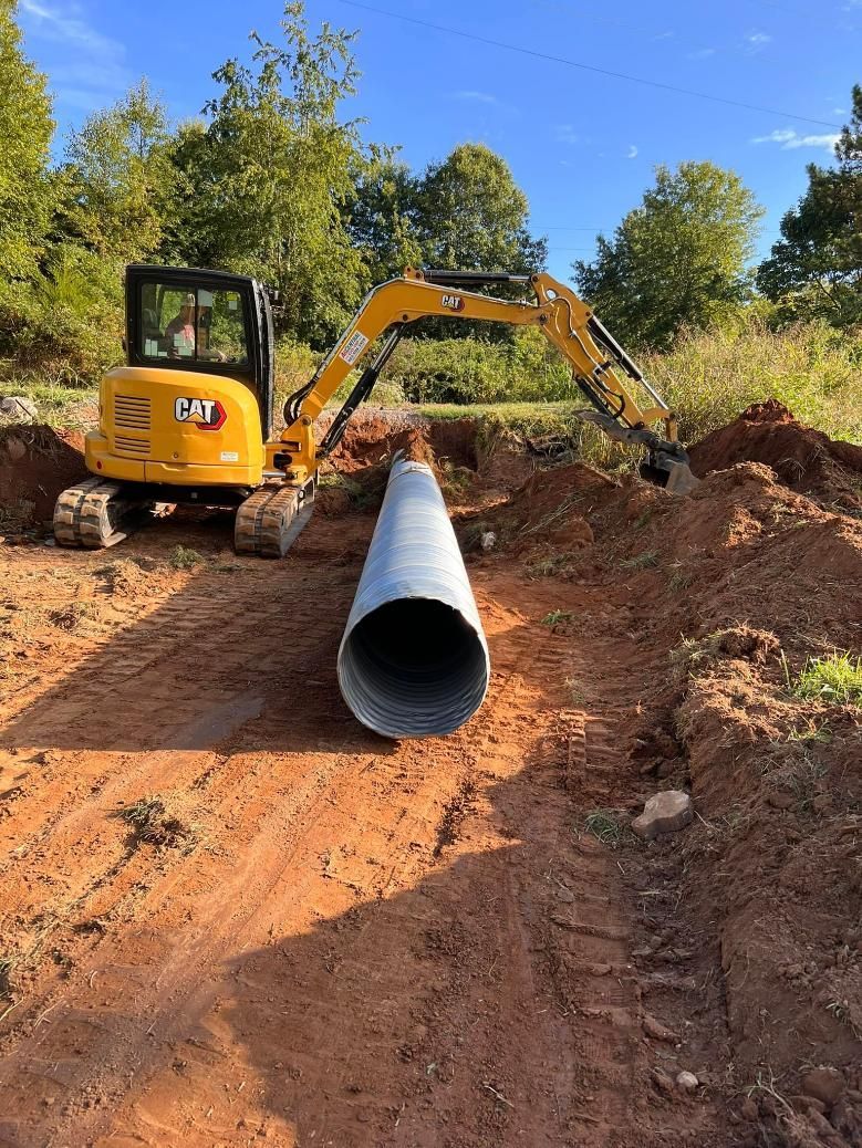 A yellow excavator is preparing to lay a large silver pipe in a dirt trench in a grassy area.