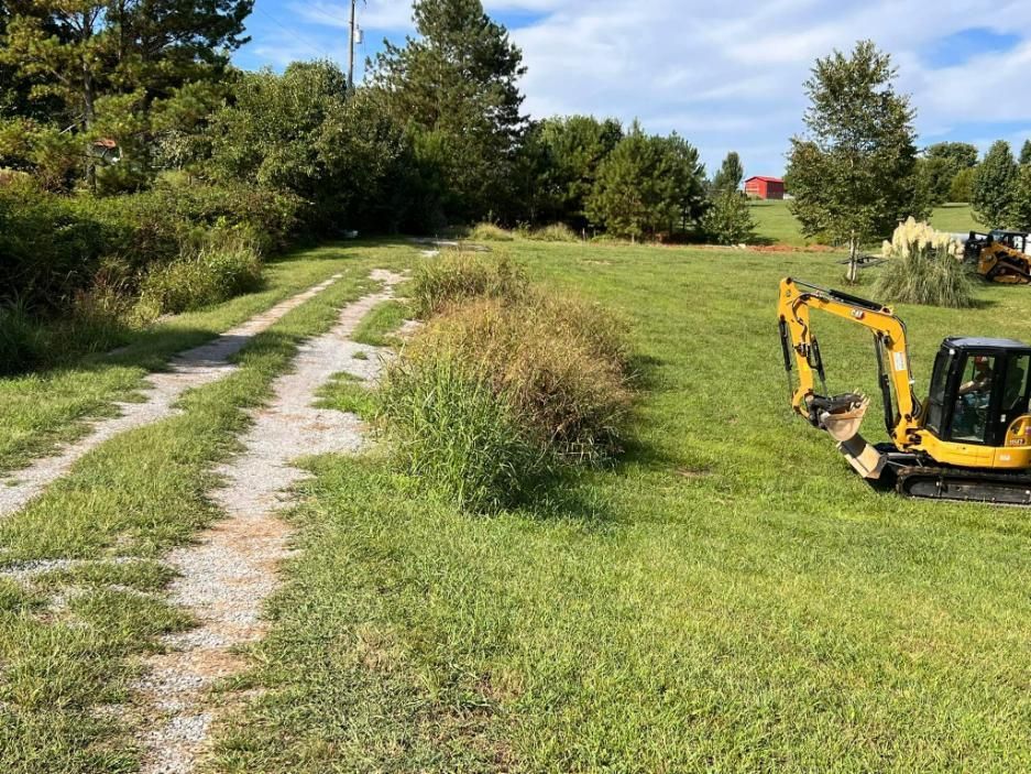 Gravel road leading through grassy field, small excavator parked nearby. Trees and blue sky in the background.