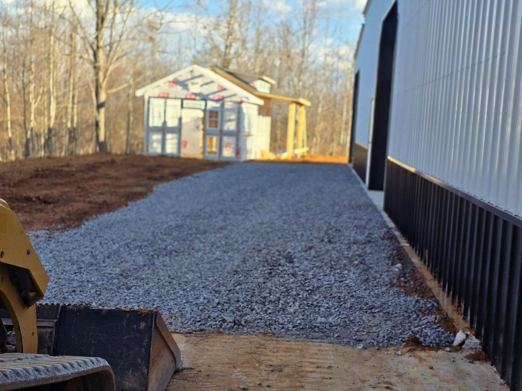 A gravel driveway leads toward a small shed, next to a metal building, with a machine in the foreground.