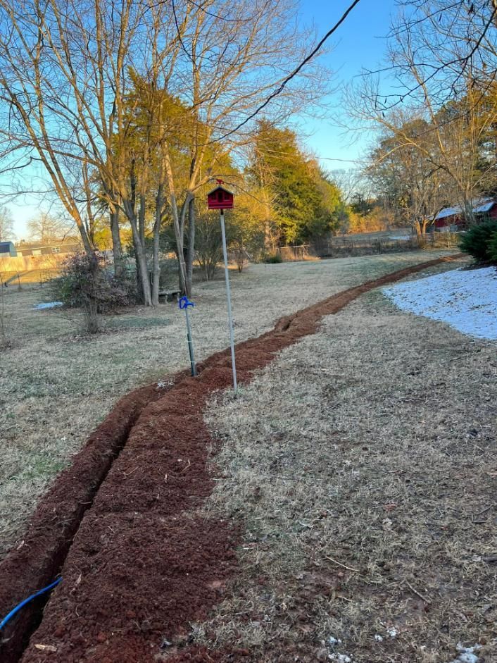 A long, shallow trench in a grassy yard. Reddish-brown soil, clear sky, a birdhouse, and sparse trees.