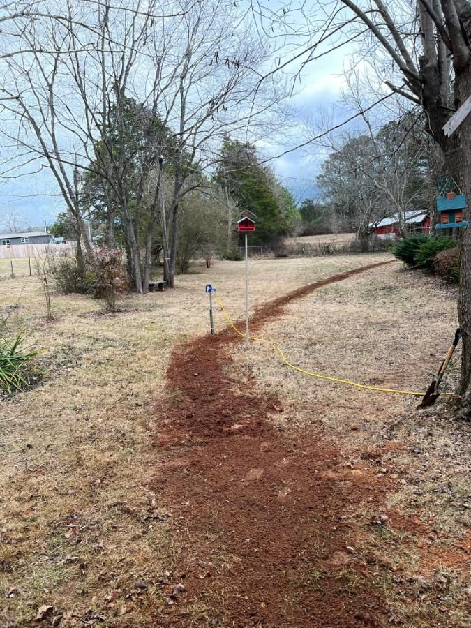 A narrow, reddish-brown dirt path winds through a grassy yard with bare trees and a cloudy sky.