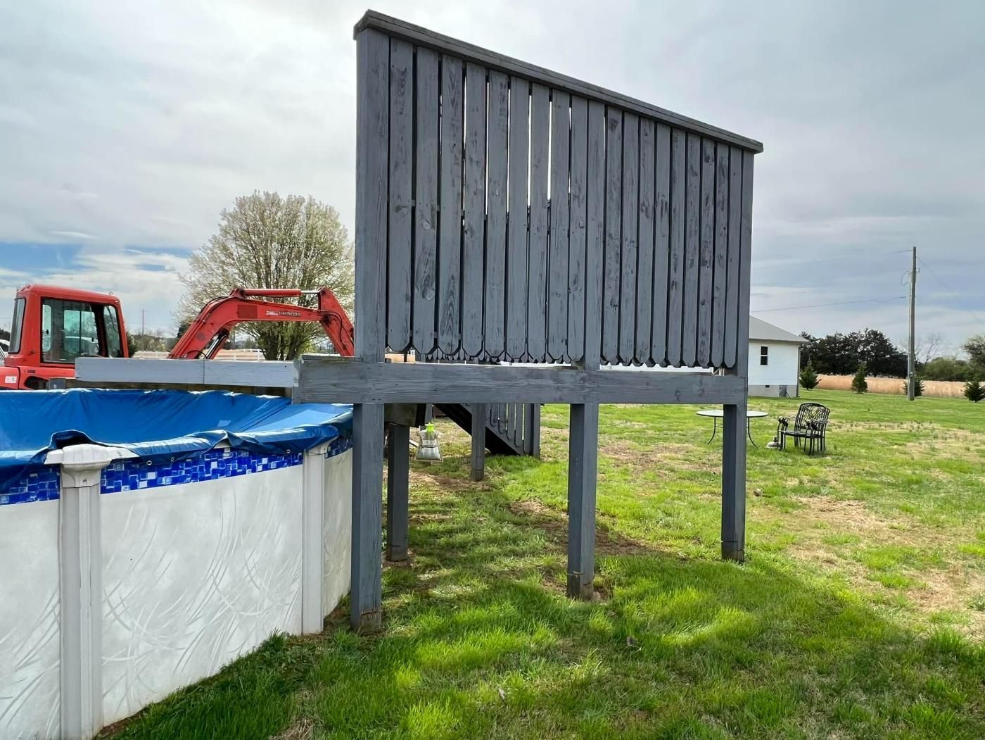 Above-ground pool with a deck and privacy screen, grey-painted. An excavator is nearby.
