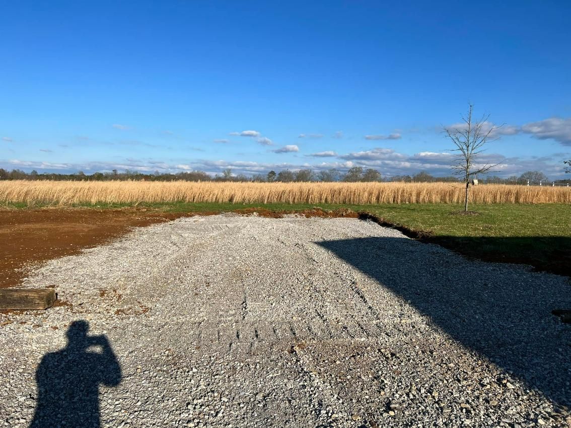 Shadow of person taking photo on gravel driveway, field and blue sky in background.