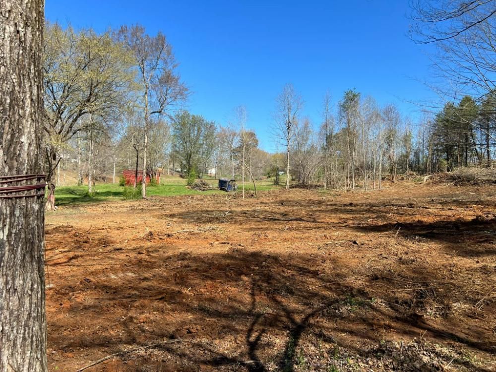 Cleared field, brown dirt, under a blue sky. Some trees, green grass, and a small building visible.