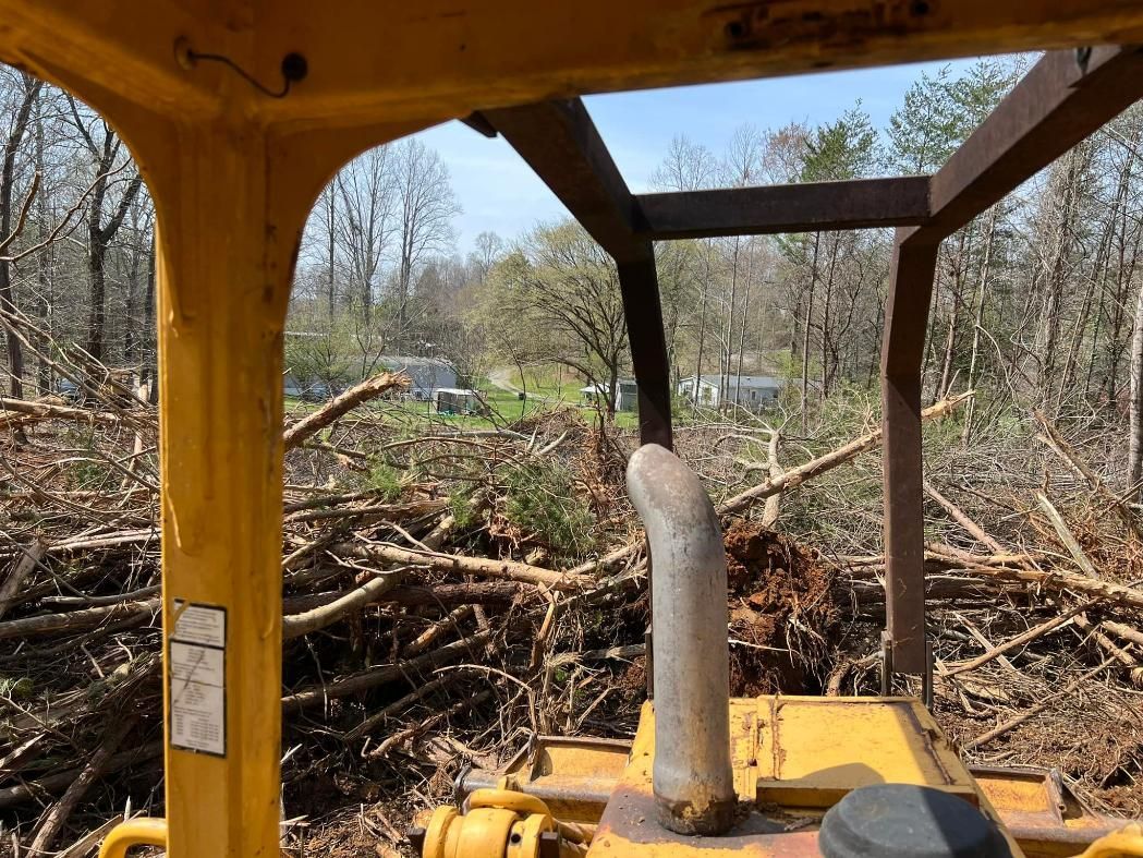 View from inside a yellow bulldozer cab, looking at felled trees and a distant house in a wooded area.