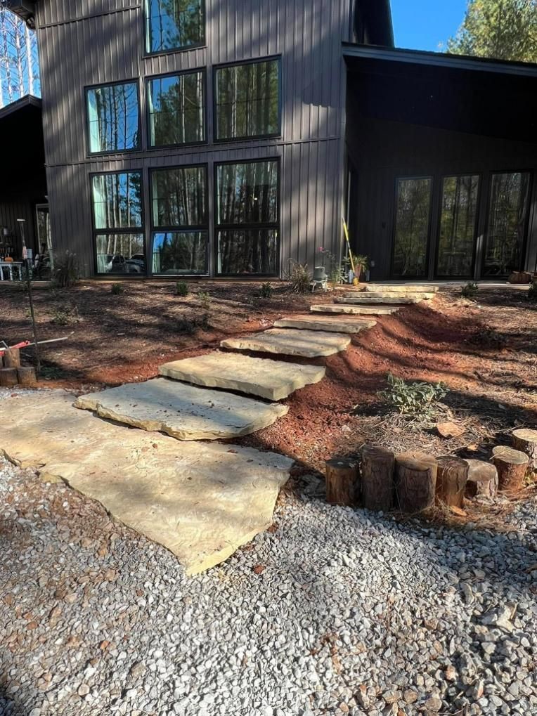 Stone steps lead to a dark-colored house with large windows. Gravel and mulch border the path.