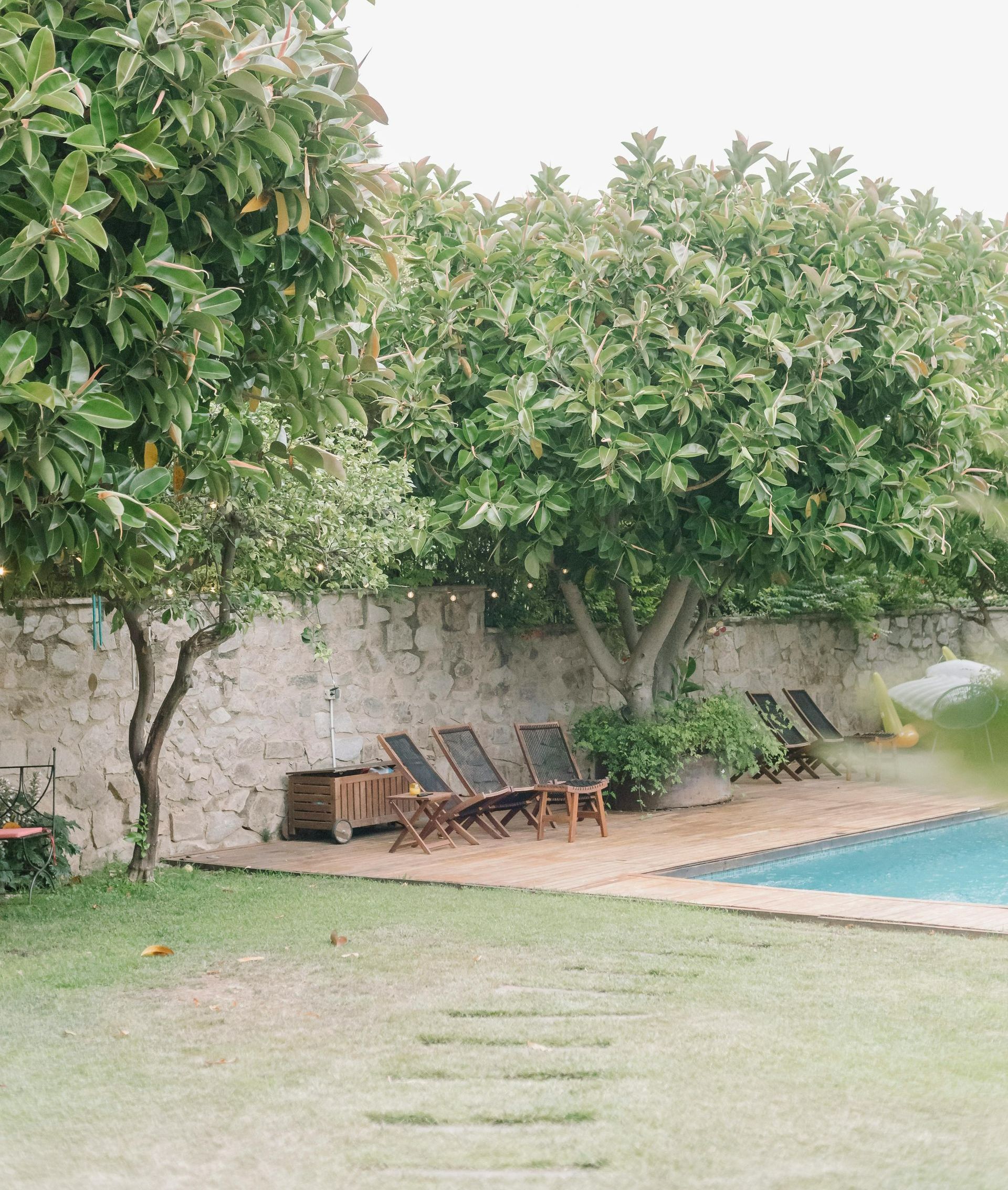 Lawn chairs by a pool near a stone wall and trees. Green grass and a cloudy sky in the background.