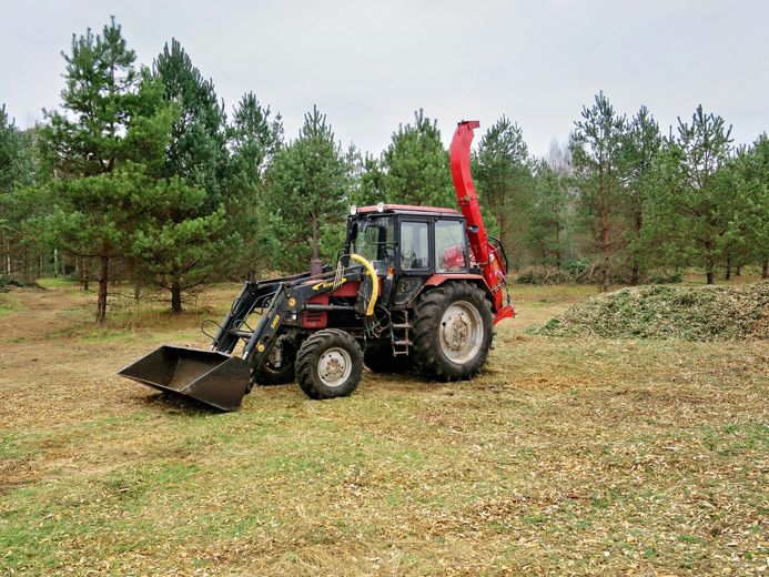Red tractor with front loader and wood chipper in a forest clearing.