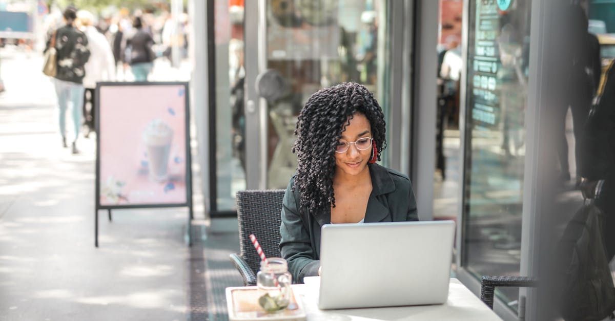 Woman with curly hair sits at a table, working on a laptop outside a cafe.