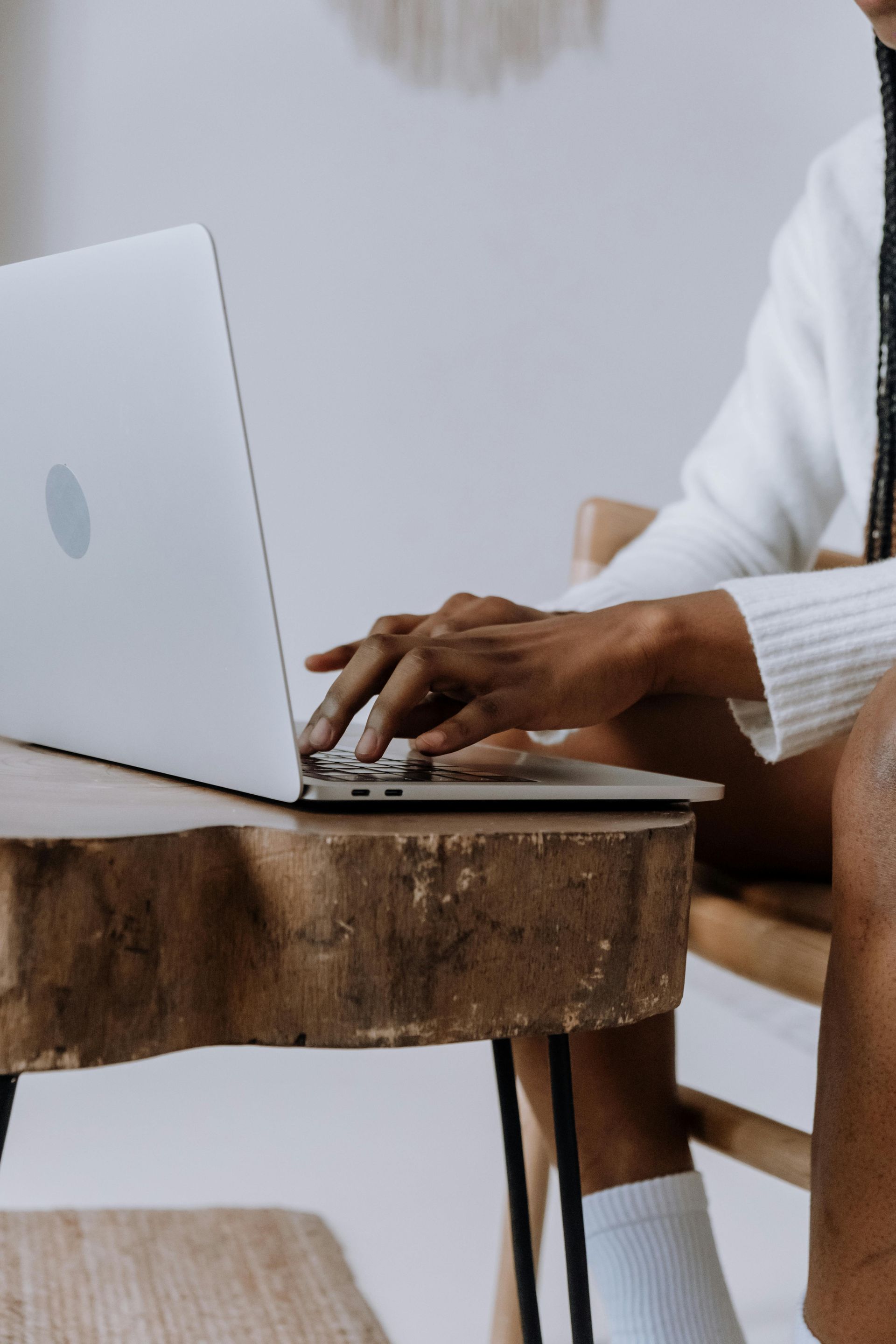 Person typing on a laptop at a wooden table, wearing a white sweater and socks.