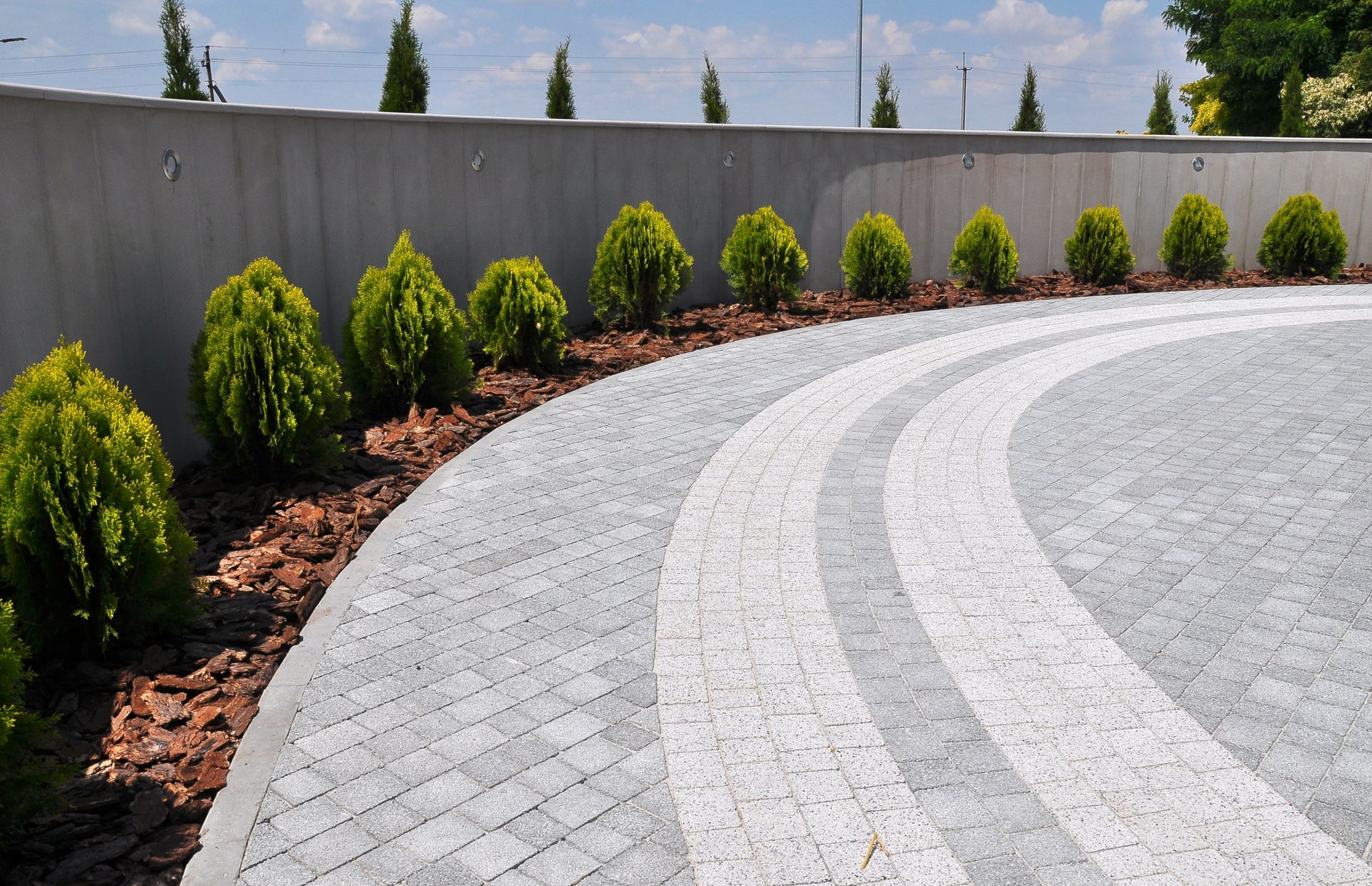 Curved stone driveway beside a row of small green shrubs and a gray wall Curved stone driveway beside a row of small green shrubs and a gray wall