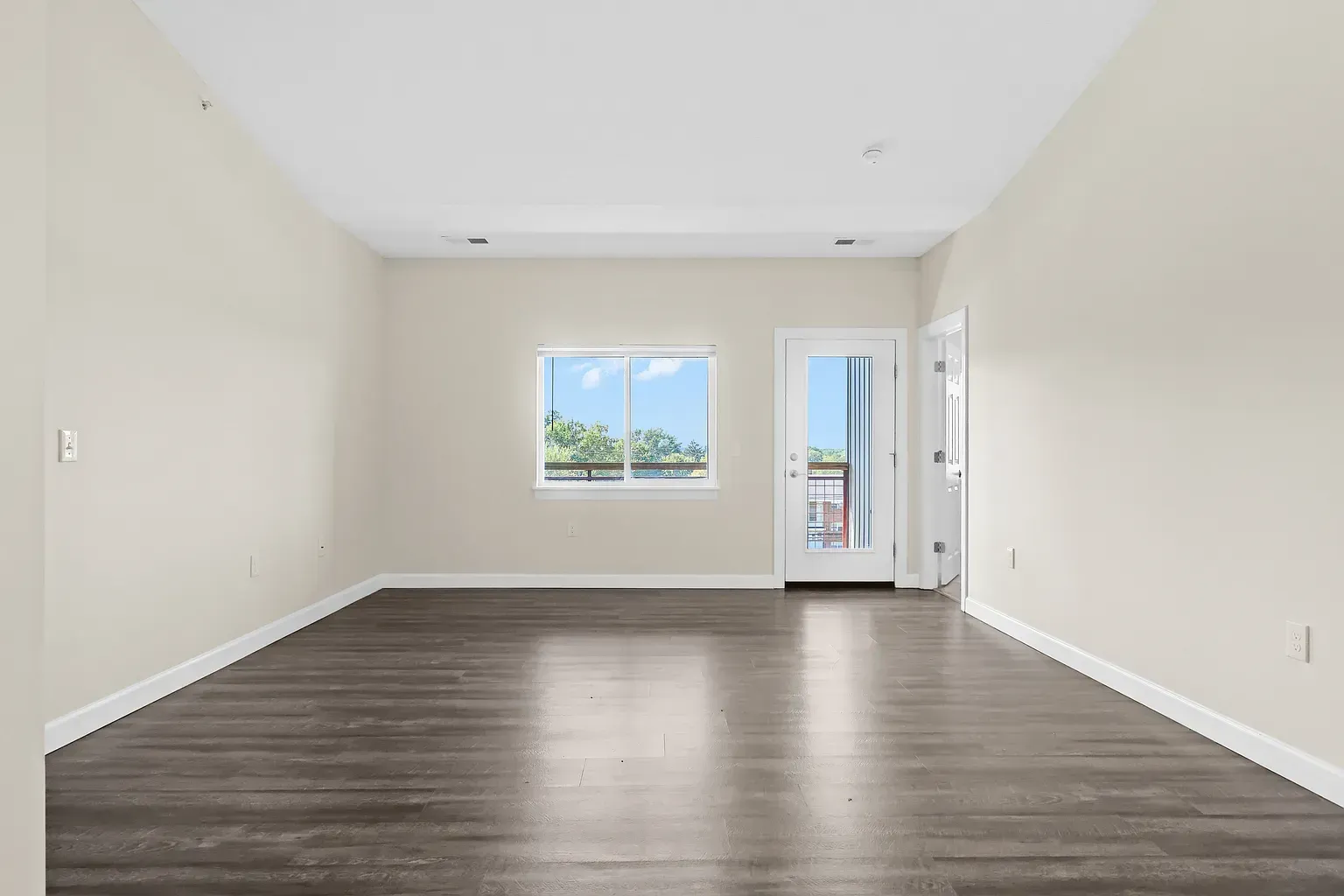 Empty living room with wood flooring, light walls, window, and door to balcony.