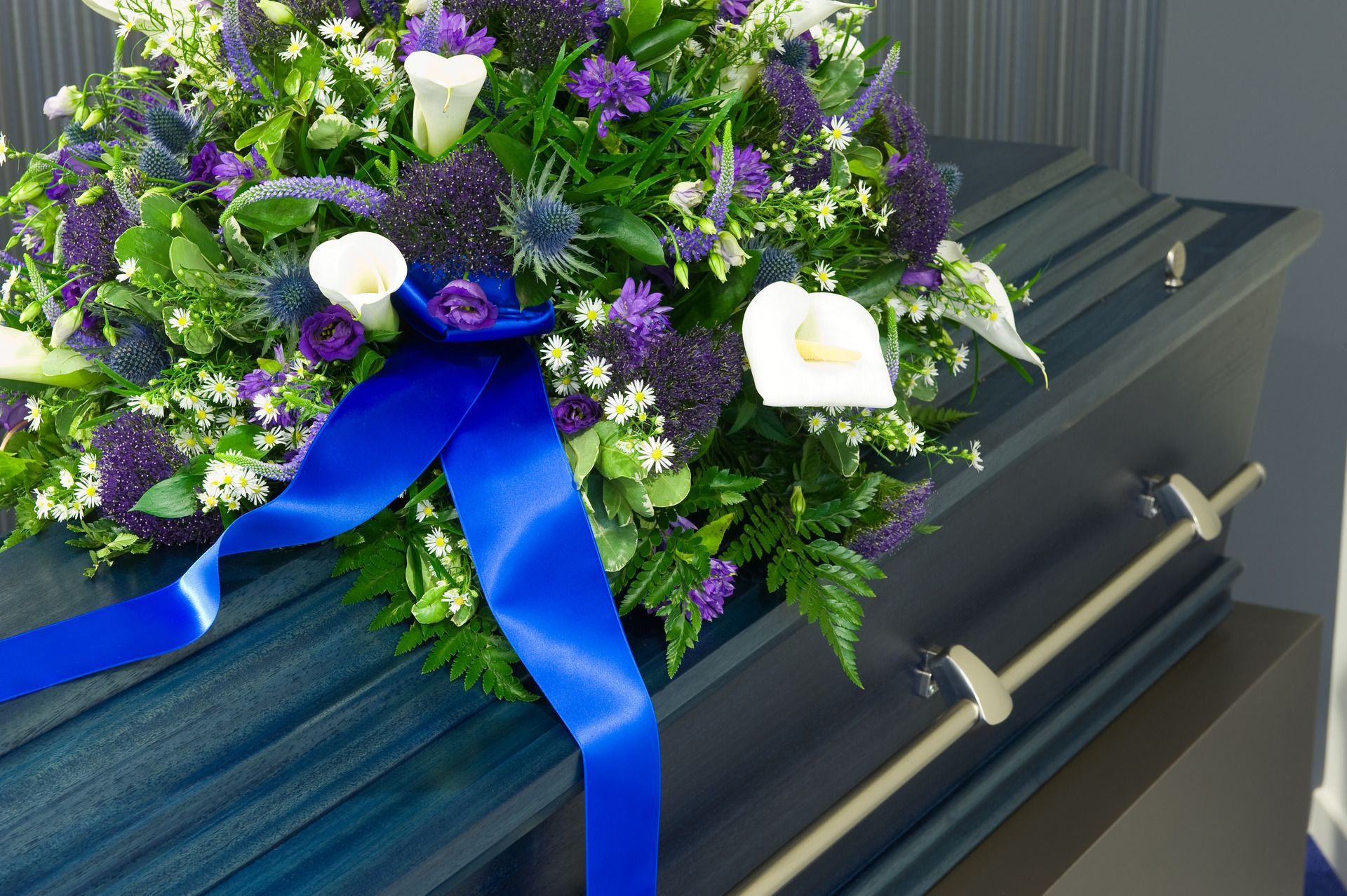 A blue casket adorned with a large floral arrangement featuring white calla lilies, blue flowers, and a bright blue ribbon.