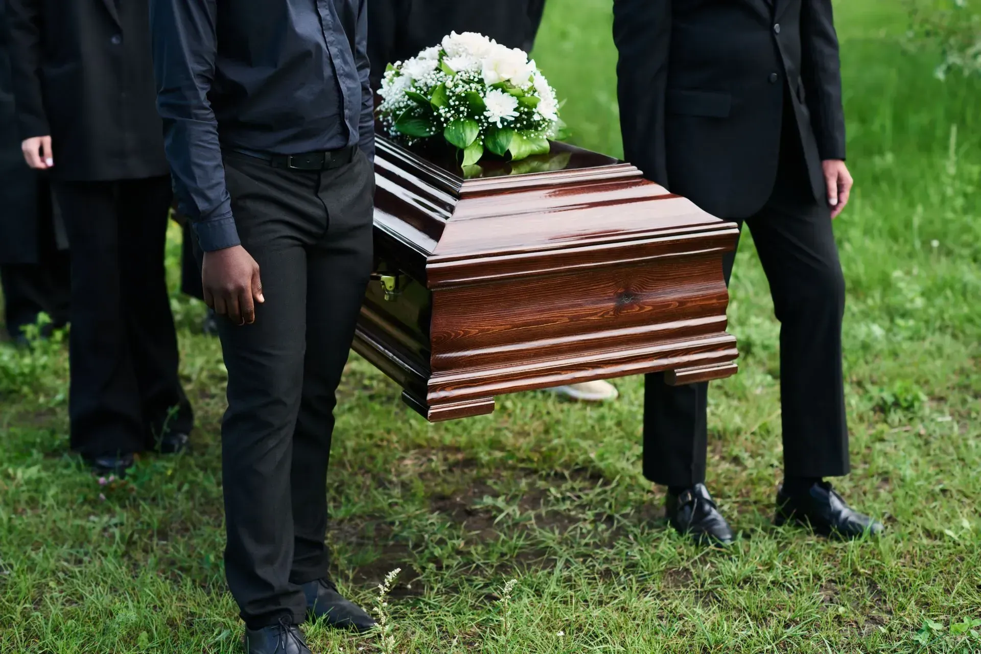 Pallbearers in dark attire carry a polished wooden casket topped with white flowers across a grassy field.