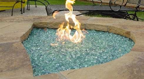 Fire pit with blue-green glass, flames, and a stone border on a patio.