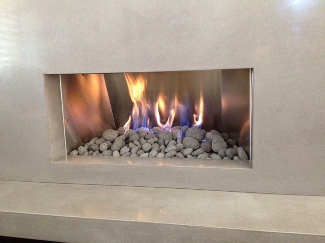 Fireplace with flames burning above gray stones in a rectangular recess within a light-colored wall.