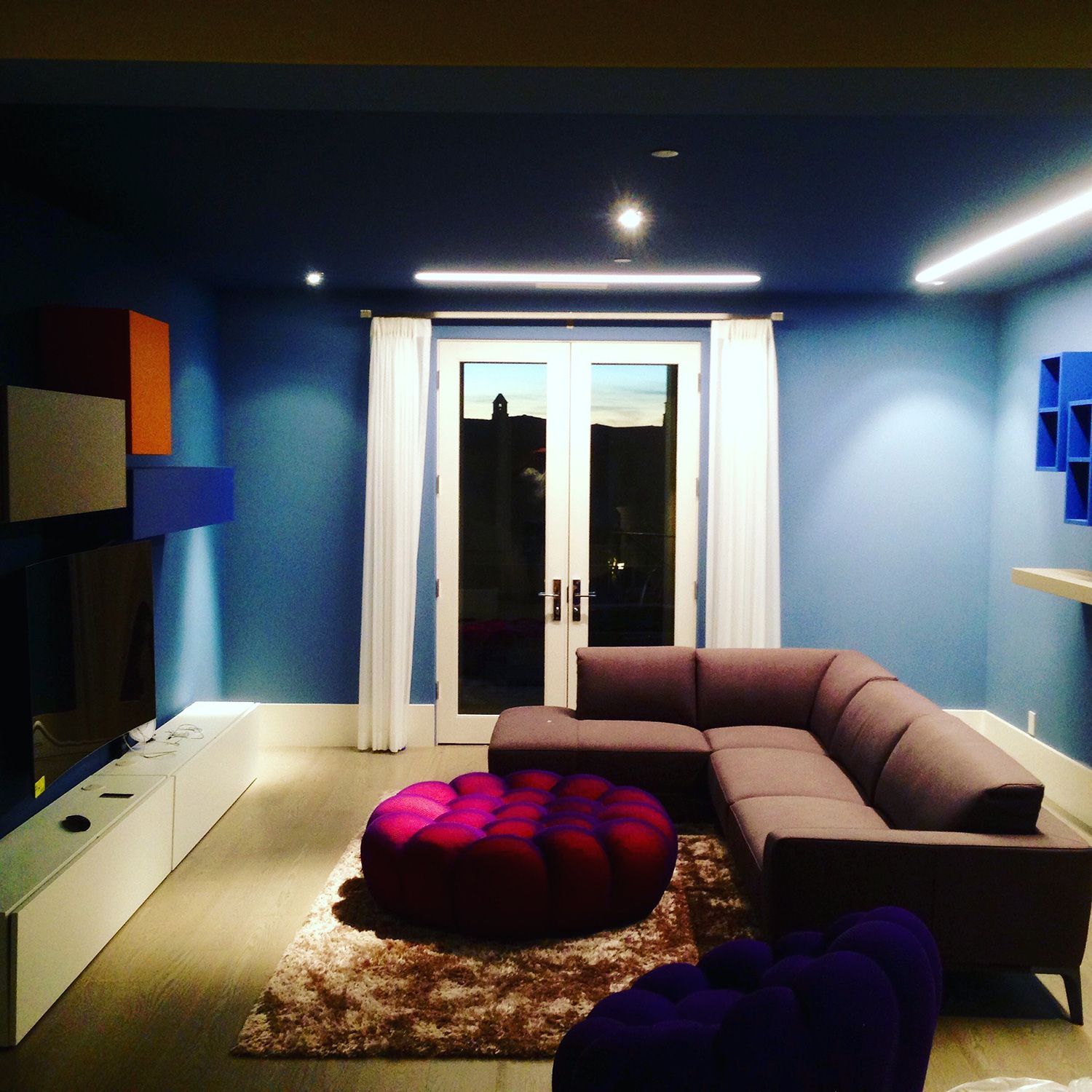 A modern living room with blue walls, gray sectional sofa, and a colorful ottoman, with double doors and a rug.
