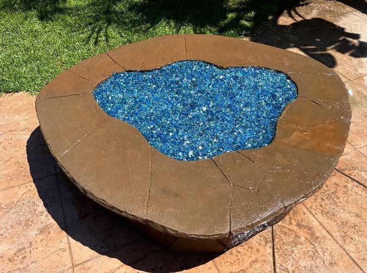 Brown stone fire pit with blue glass, on a patio, in sunlight.