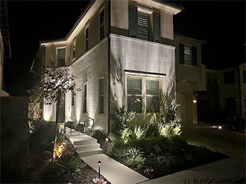 Two-story house with outdoor lighting at night, illuminating the facade, pathway, and landscaping.