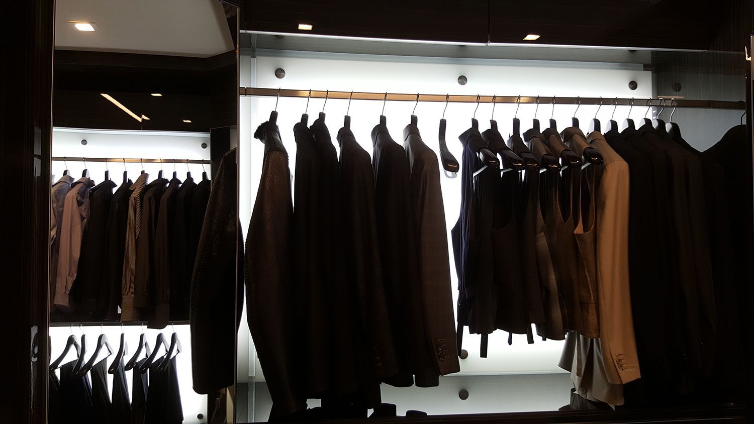 Closet with suits and jackets hanging on display. Dark interior, bright backlighting illuminates the clothing.