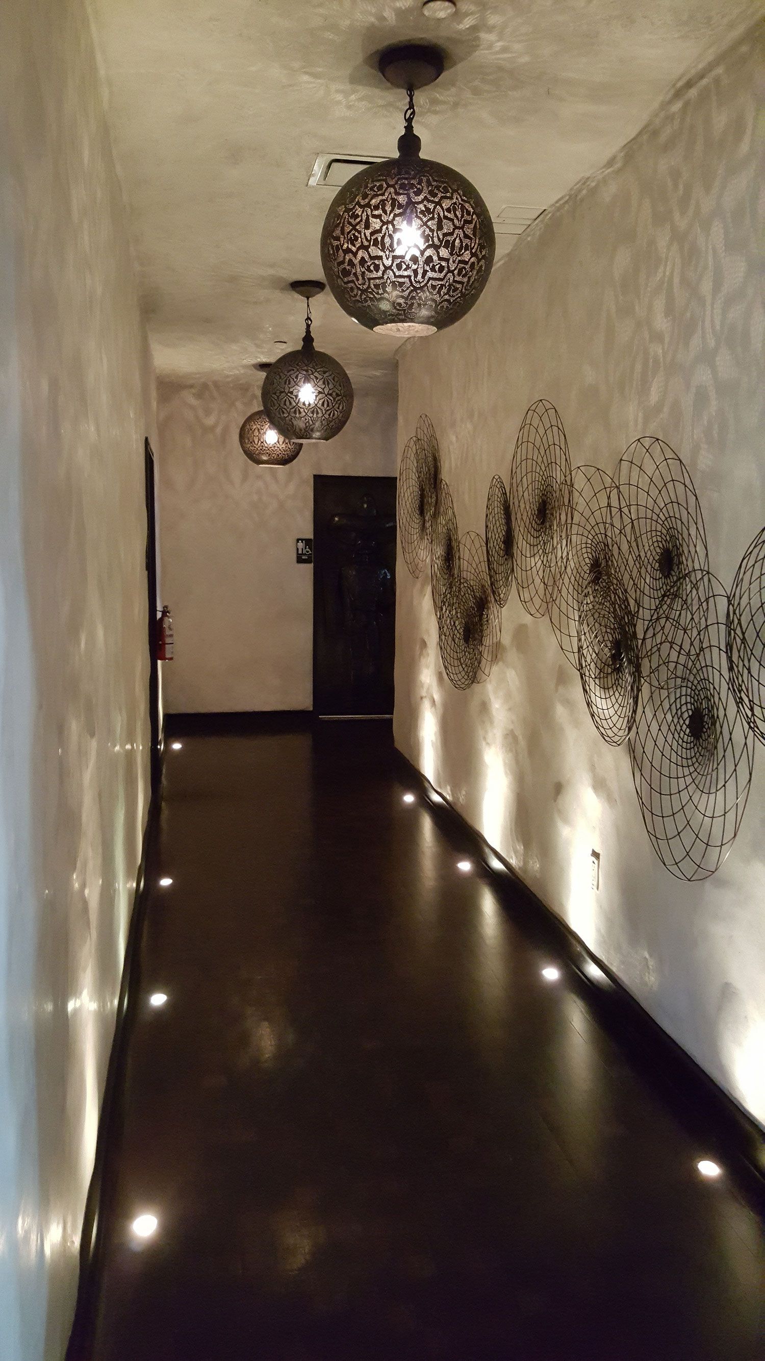 Narrow hallway with dark floor, lit by overhead globe lamps and floor lights; decorative wall art.