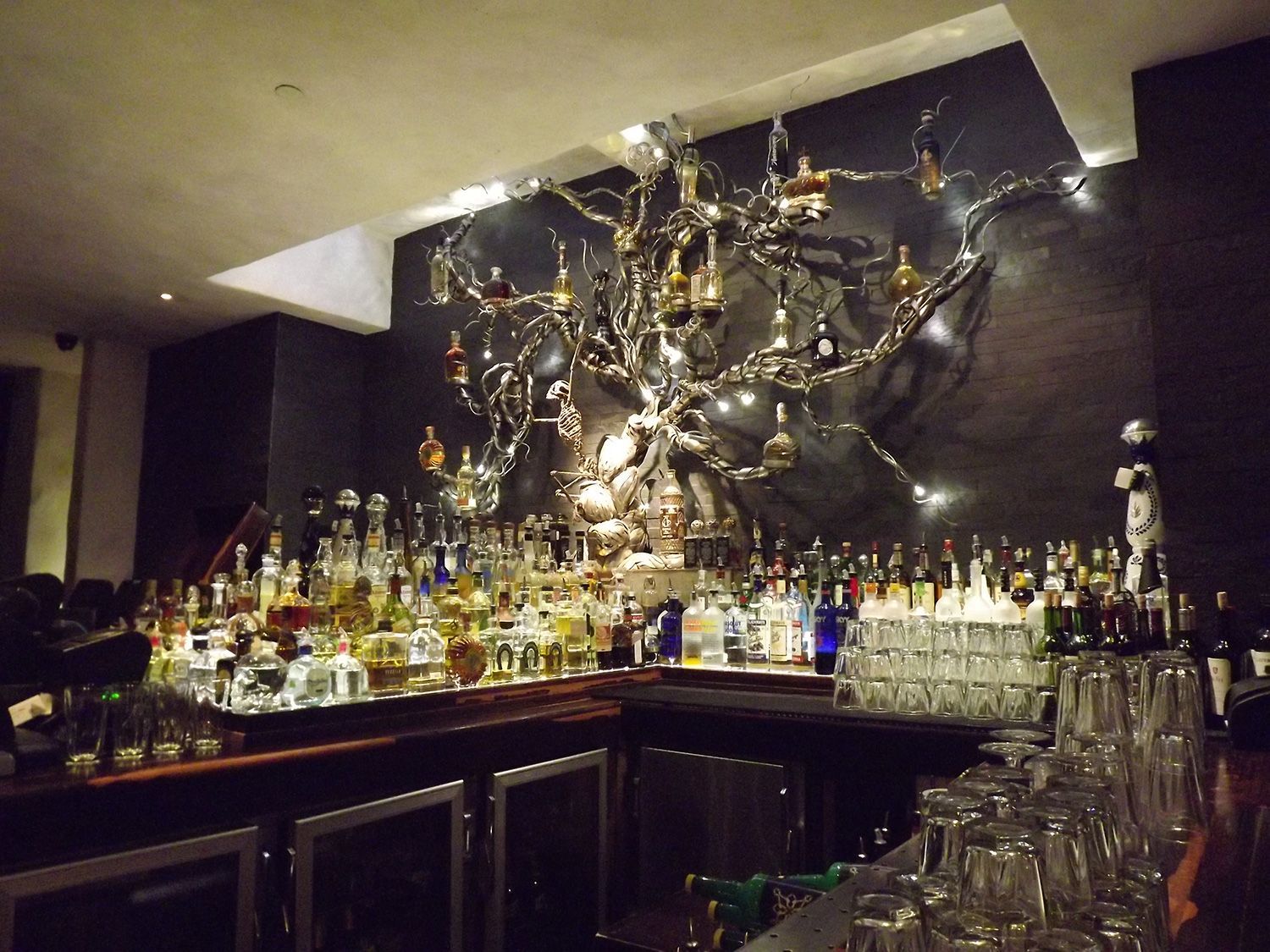 Bar with bottles, glasses, and a large metal tree sculpture on the back wall.