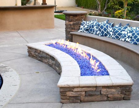Curved fire pit with blue glass, stone base, and built-in bench with blue patterned pillows. Outdoor patio setting.