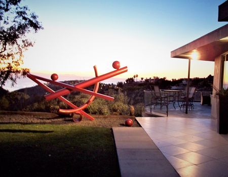 Red abstract sculpture on a lawn beside a modern house patio, overlooking a hillside at sunset.