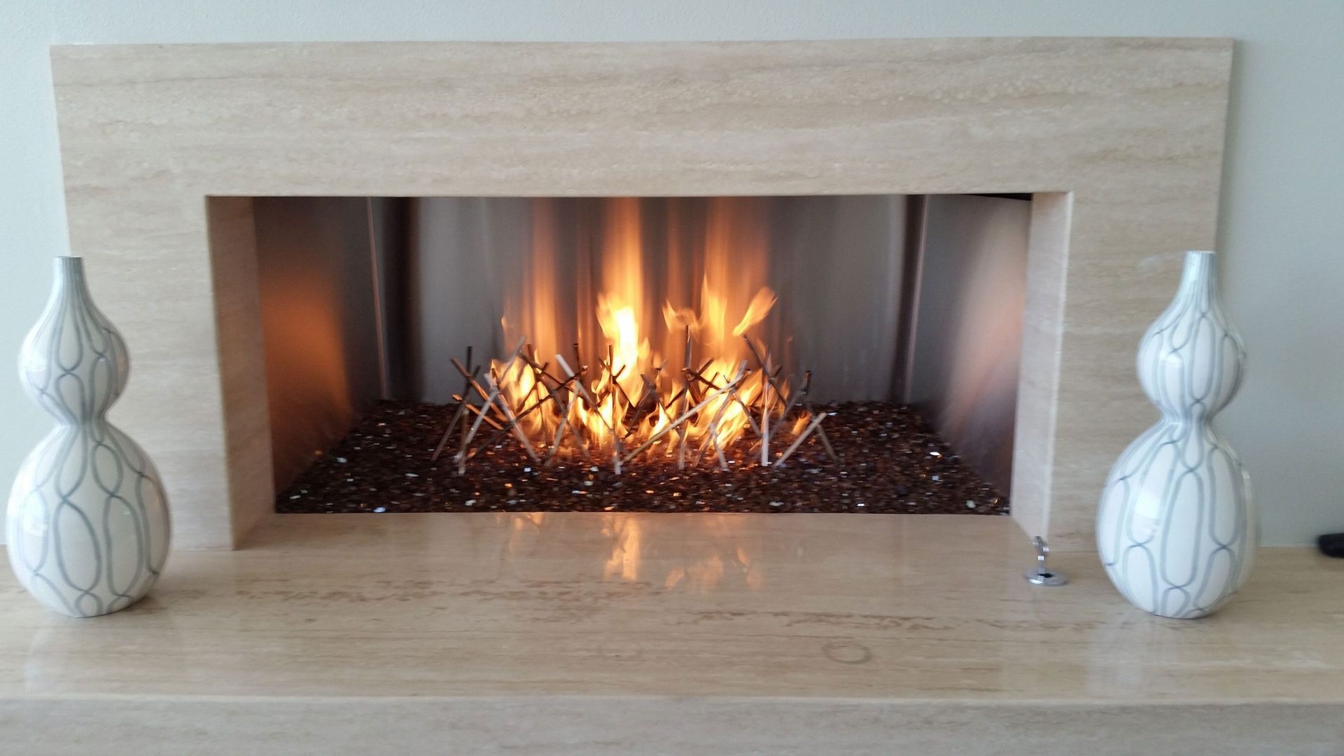 Fireplace with flames, beige mantle, glass rocks, and two decorative white vases.