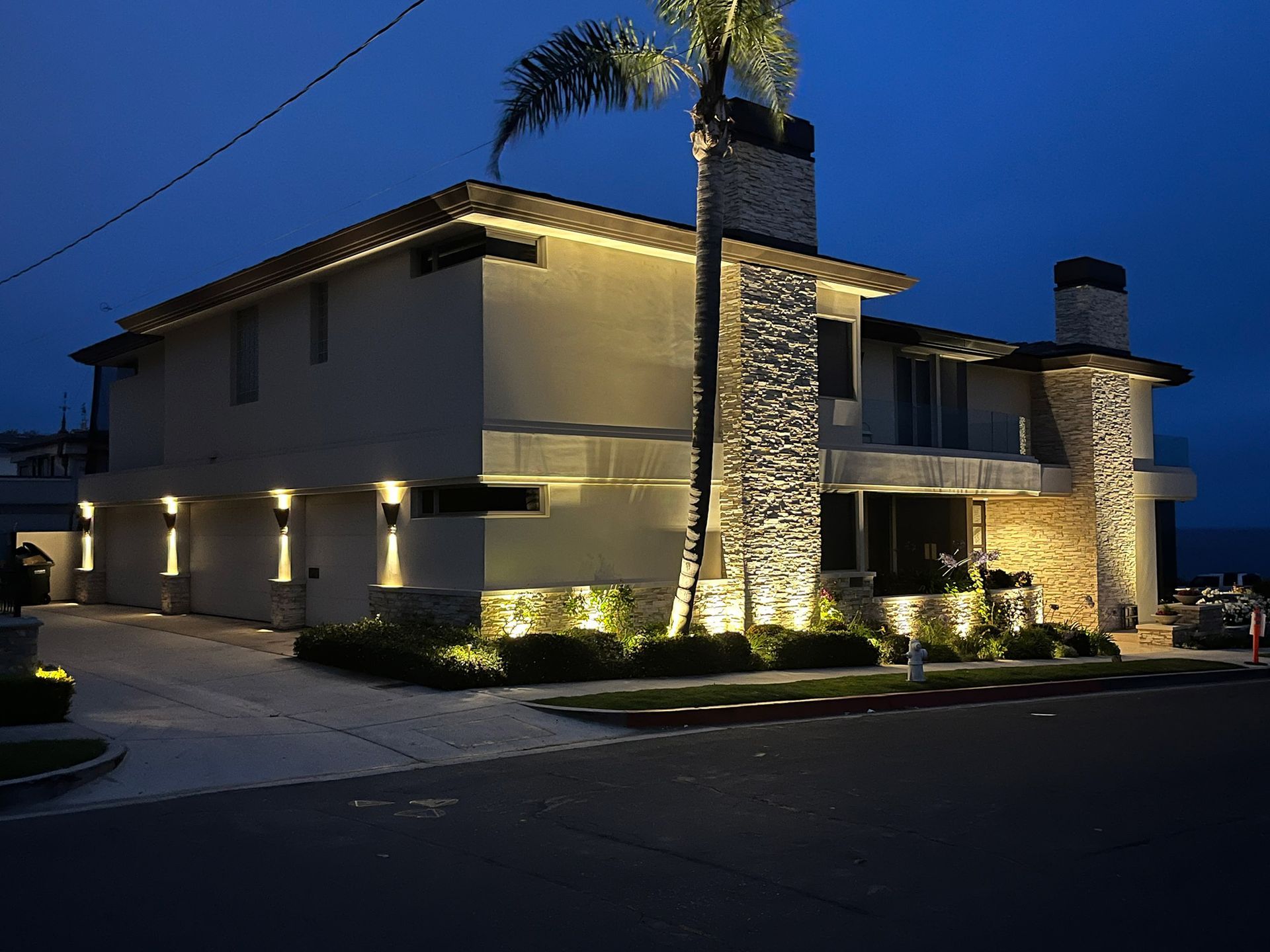 Lit-up house at dusk with stonework chimney, palm tree, and driveway.