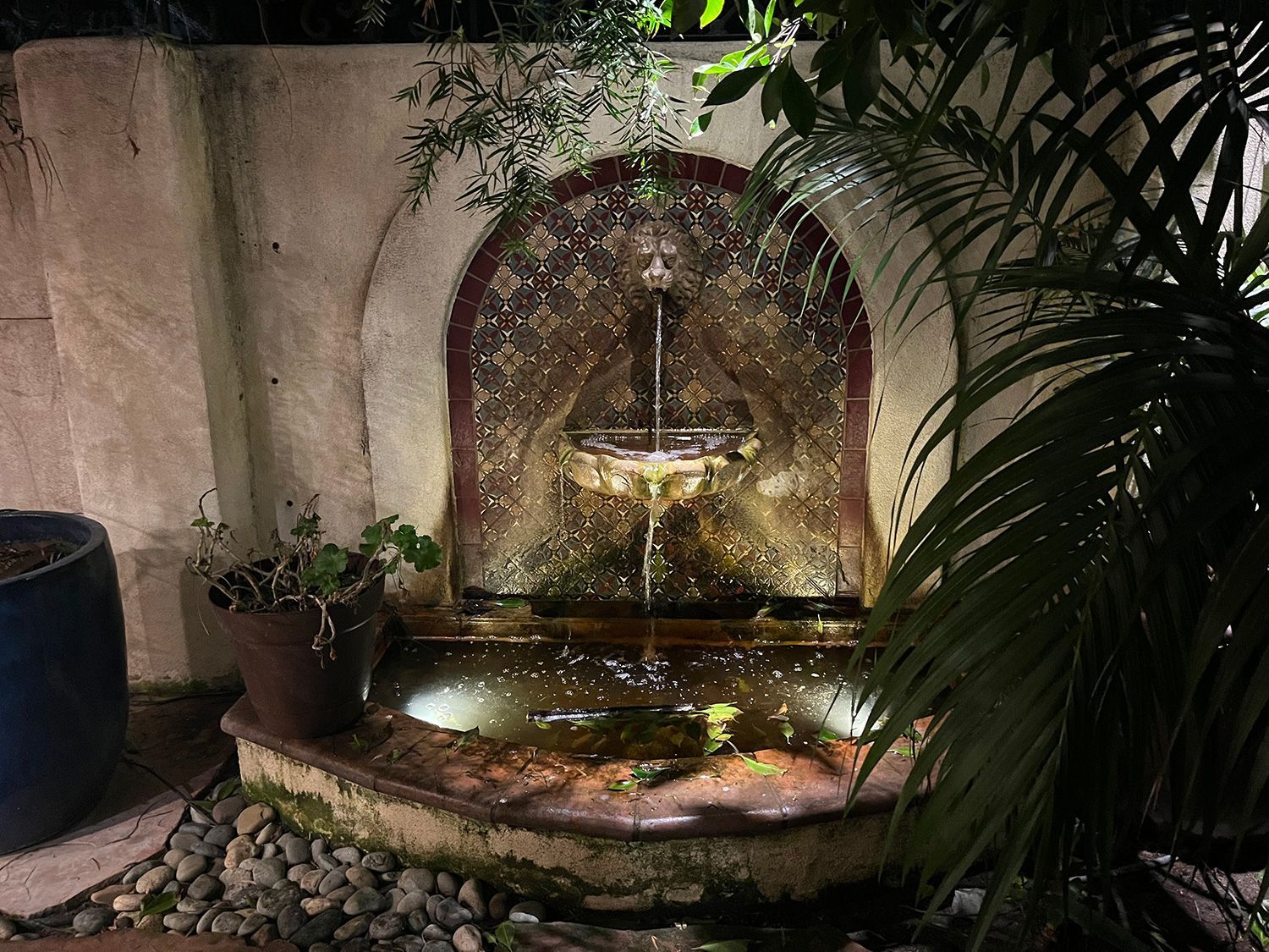 Water fountain at night, illuminated.  Dark background, arched mosaic design, water flowing, and surrounding plants.