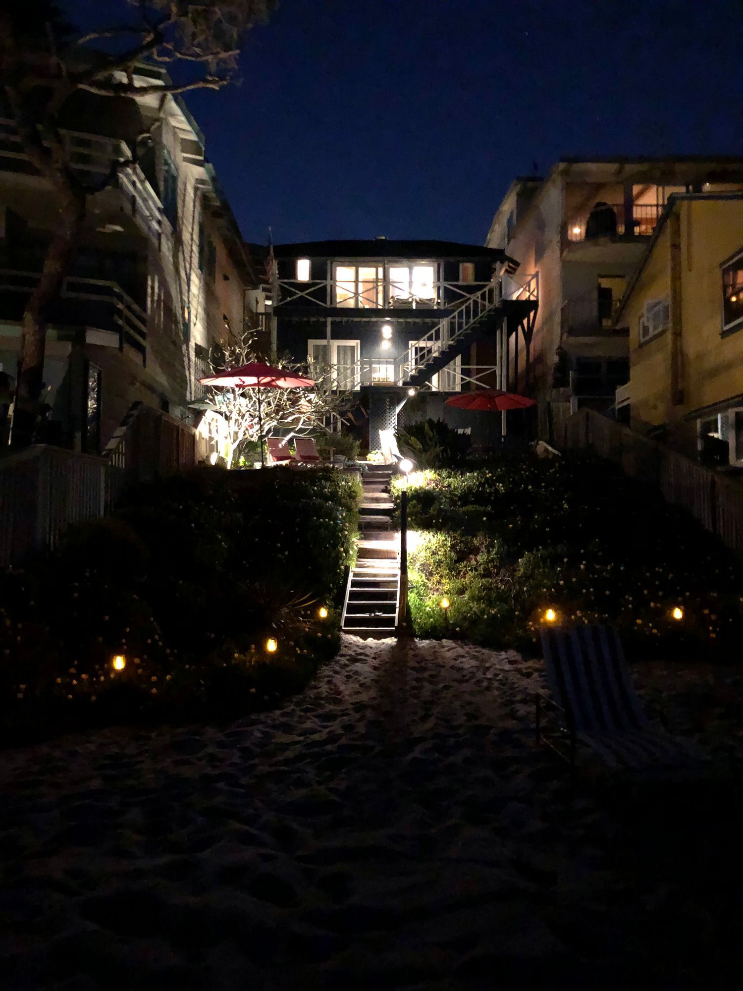 Nighttime beach scene. Stairs lead to a house lit from within, with string lights and red umbrellas visible.