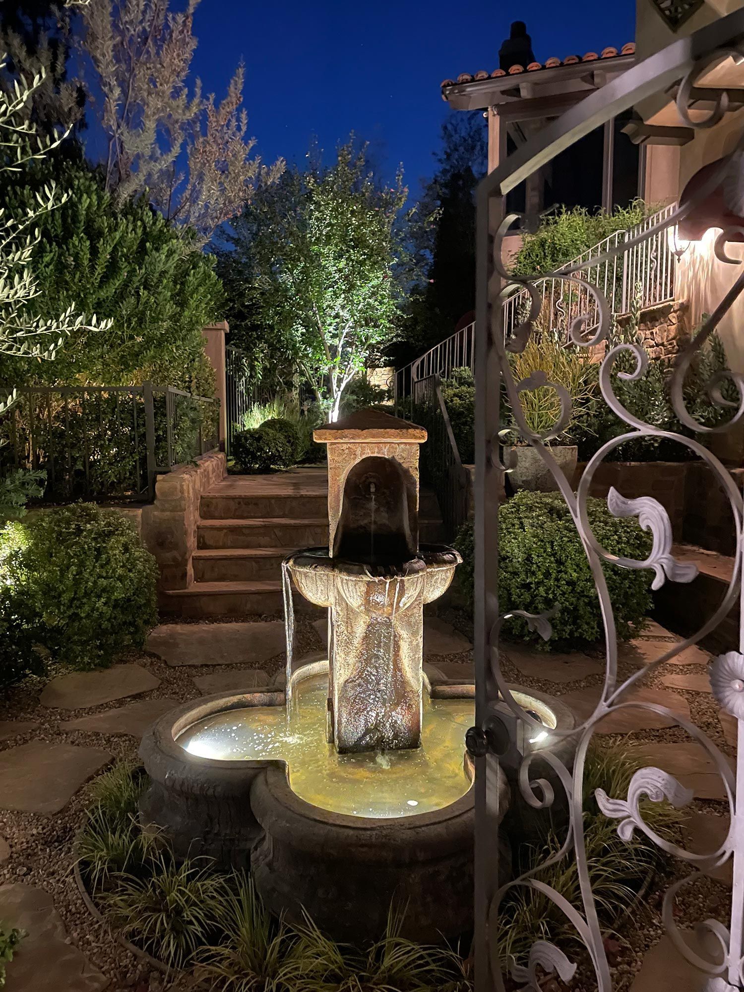 Stone fountain in a garden at night, illuminated by lights; staircase and wrought iron gate are visible.
