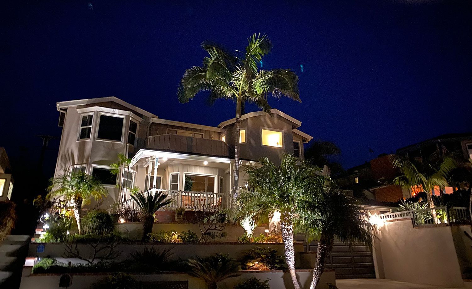 Lit-up two-story house at night, with palm trees, pathway lights, and a dark blue sky.