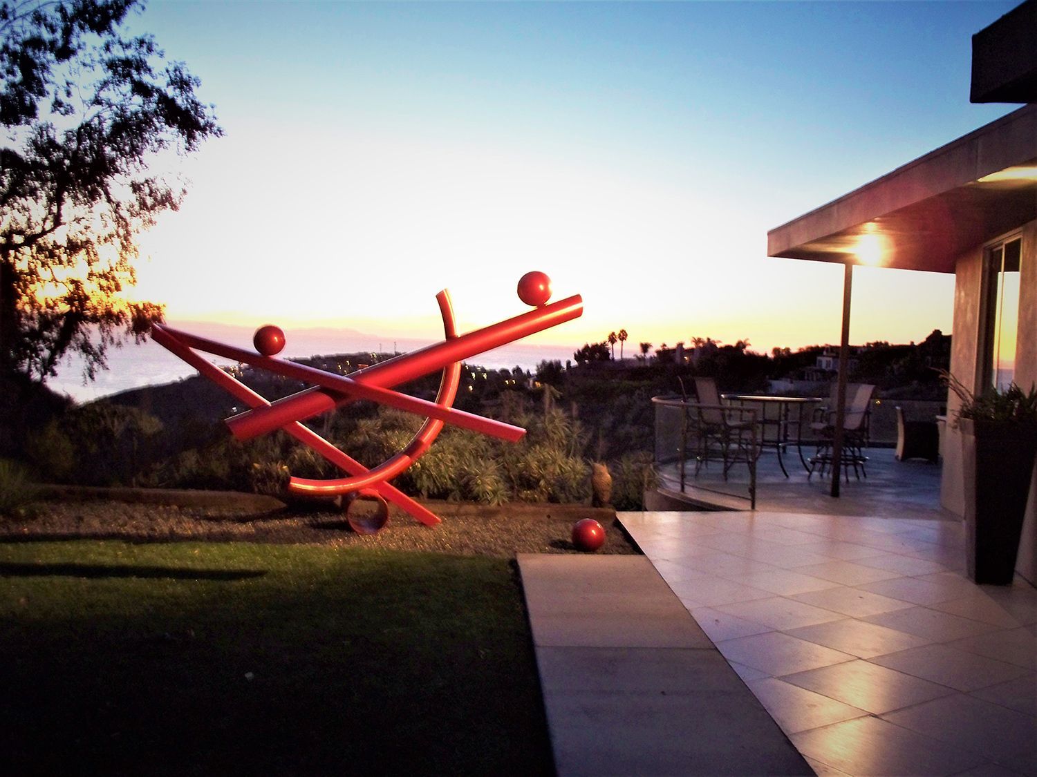 Red abstract sculpture on a lawn overlooking a sunset, next to a house patio.