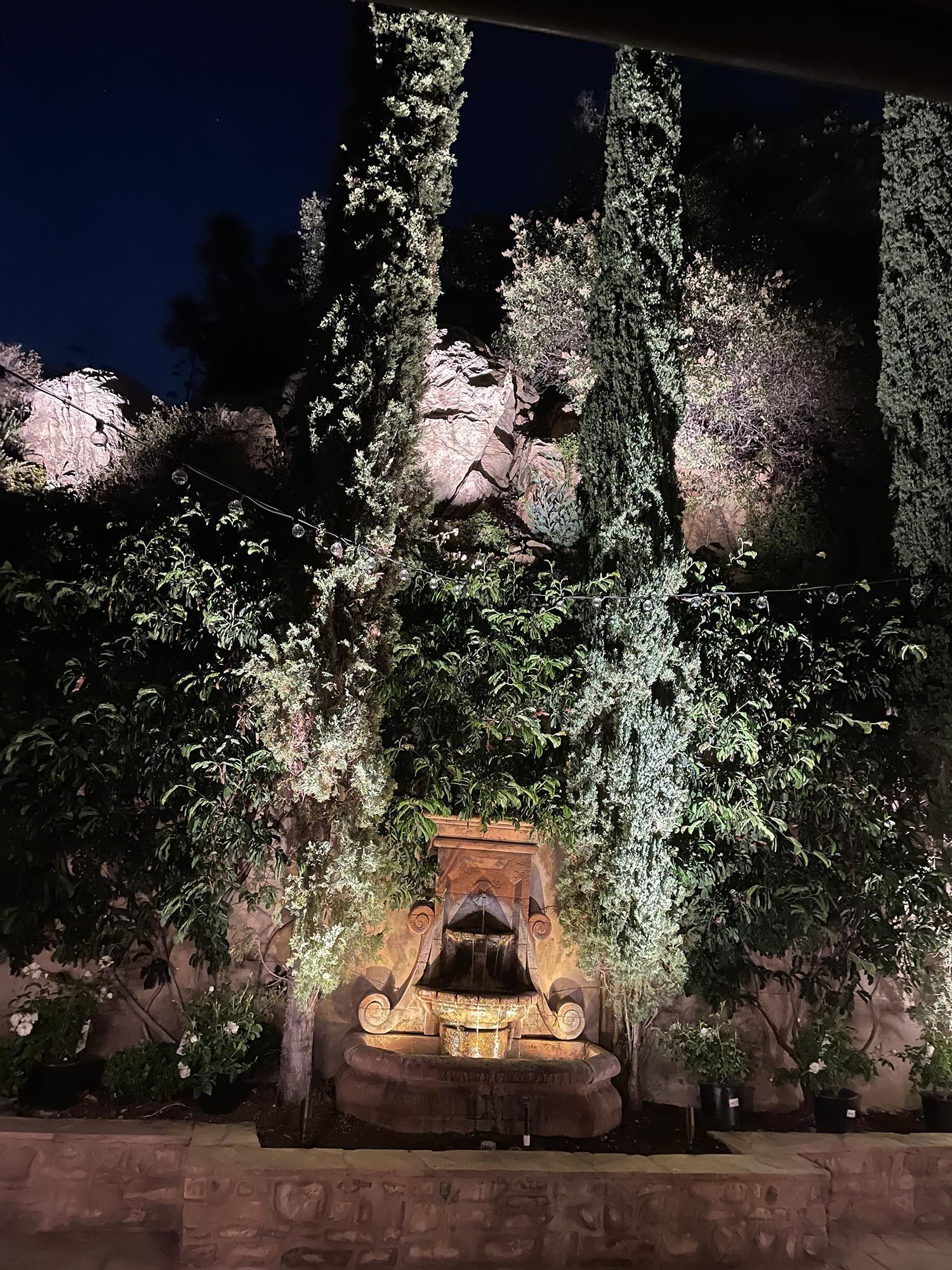 Fountain at night, lit by spotlights. Stone structure with water flowing, surrounded by greenery.