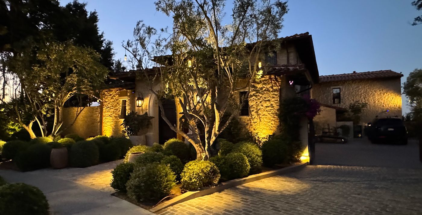 Stone house with nighttime landscape lighting; trees, bushes, and driveway illuminated.