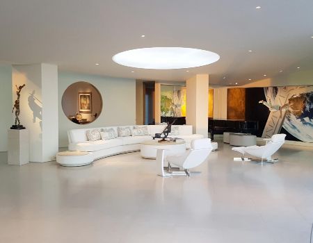 Spacious, modern living room with white curved sofa, armchairs, art, and skylight in ceiling.
