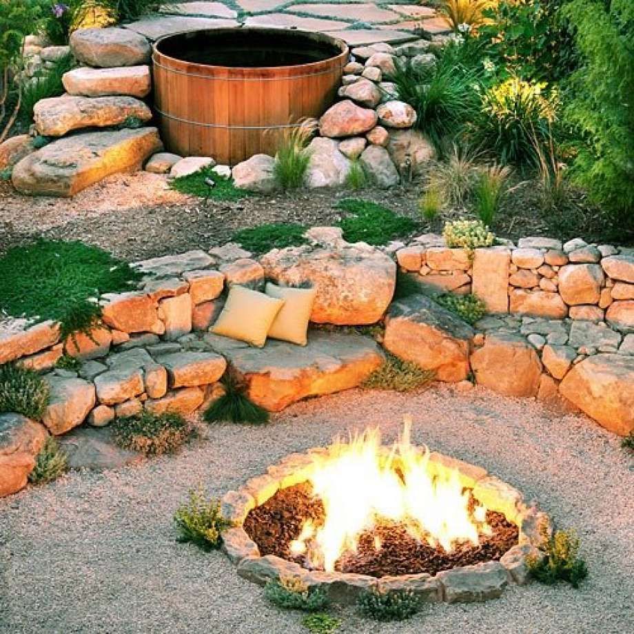 A wooden hot tub and stone seating surround a fire pit in a landscaped outdoor space.