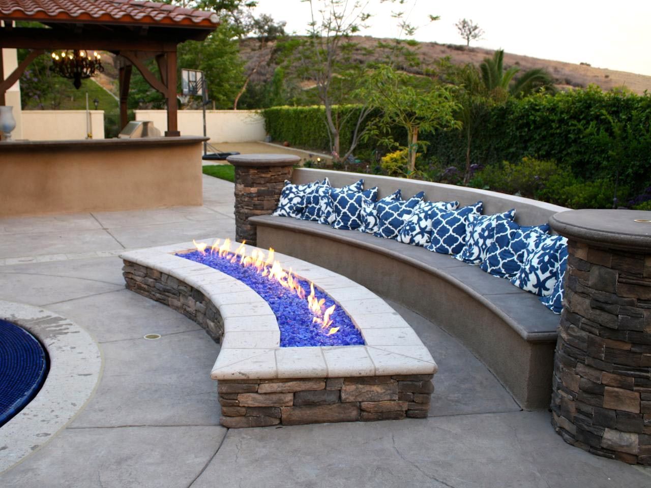 Stone fire pit with blue flames and cushioned seating in a backyard setting.