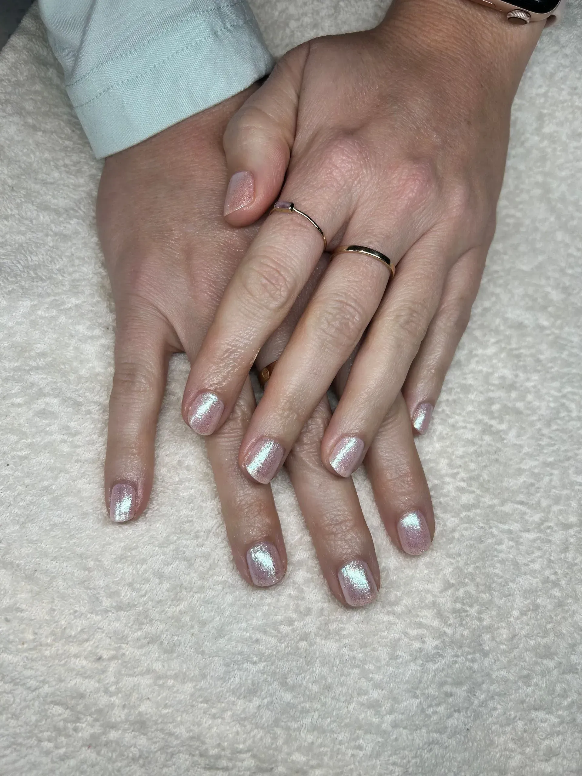Hands with shimmering light pink nail polish, wearing rings, resting on a white surface.