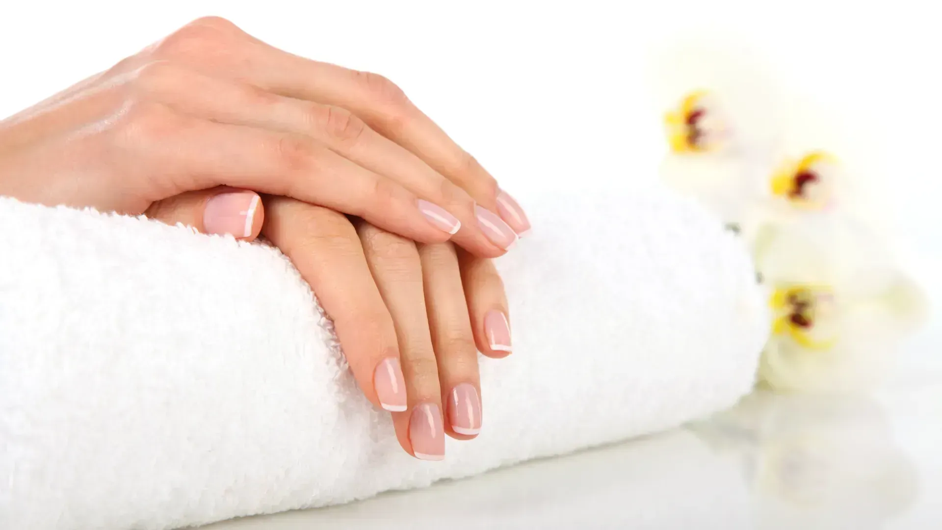 Hands with French manicure resting on a white rolled towel, with white flowers.