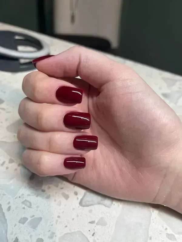 Hand with square-shaped nails painted a dark red color resting on a textured white surface.