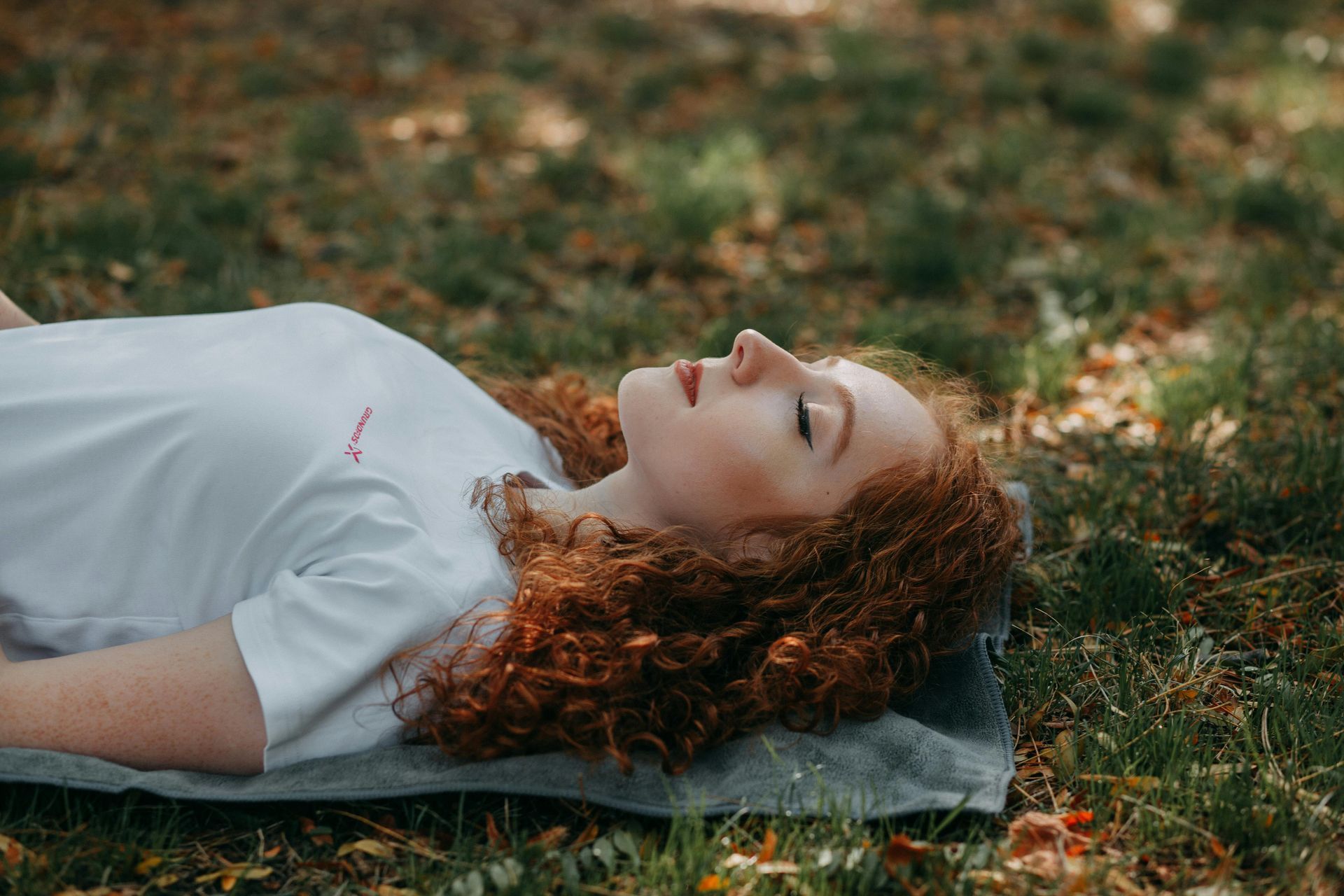 Woman with red curly hair lying on a blanket in the grass with eyes closed, lit by sunlight.