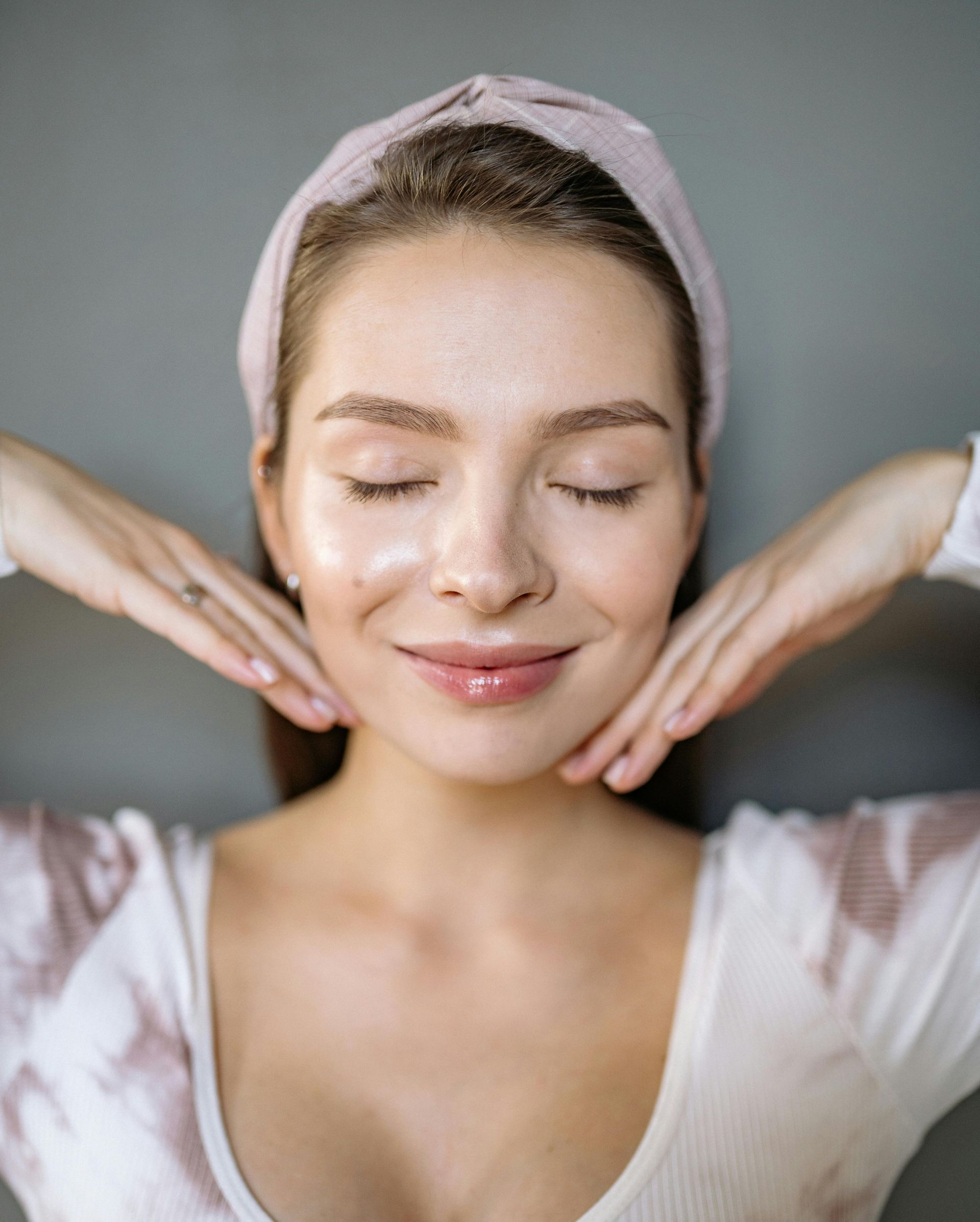 Woman with eyes closed, hands on cheeks, wearing a headband and white top, smiling.